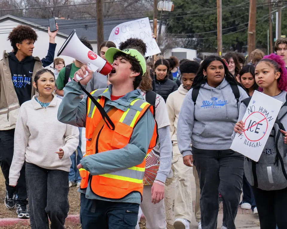 Reese, a white nonbinary person, holds a megaphone at an anti-ICE rally at McCallum High School while a crowd of students walks behind them.